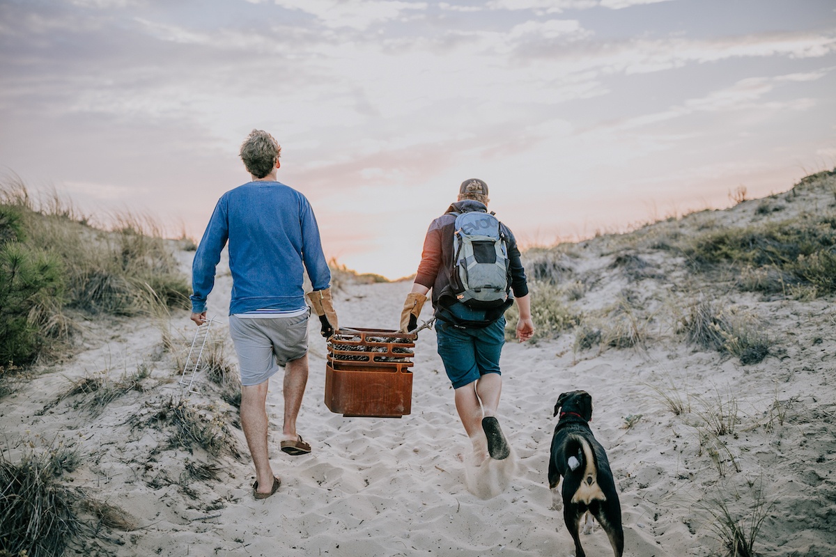 Zwei Männer tragen einen Feuerkorb über Dünen, ihnen folgt ein Hund