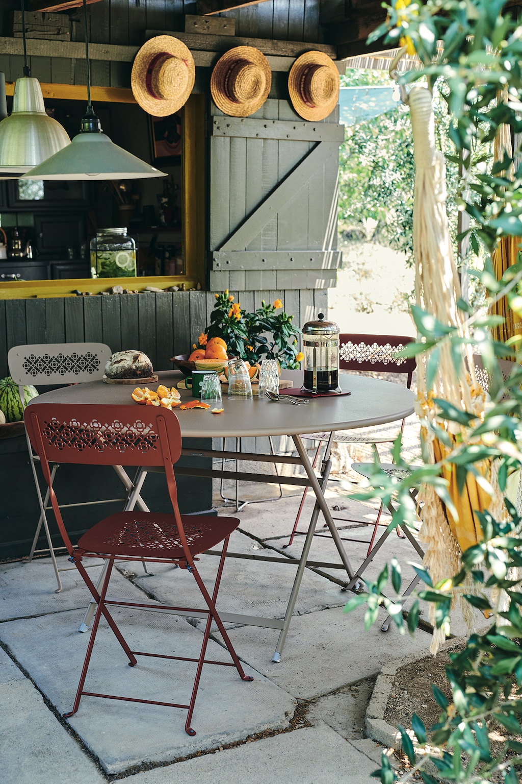 Fermob Caractère Gartentisch auf einer kleinen Terrasse. An einem Fensterladen hängen drei Strohhüte.