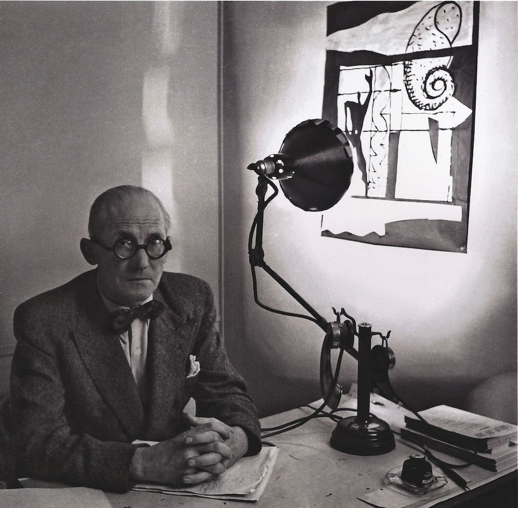 Le Corbusier an einem Schreibtisch. Rechts strahlt eine zur Wand ausgerichtete Lampe Gras ein abstraktes Bild an. 