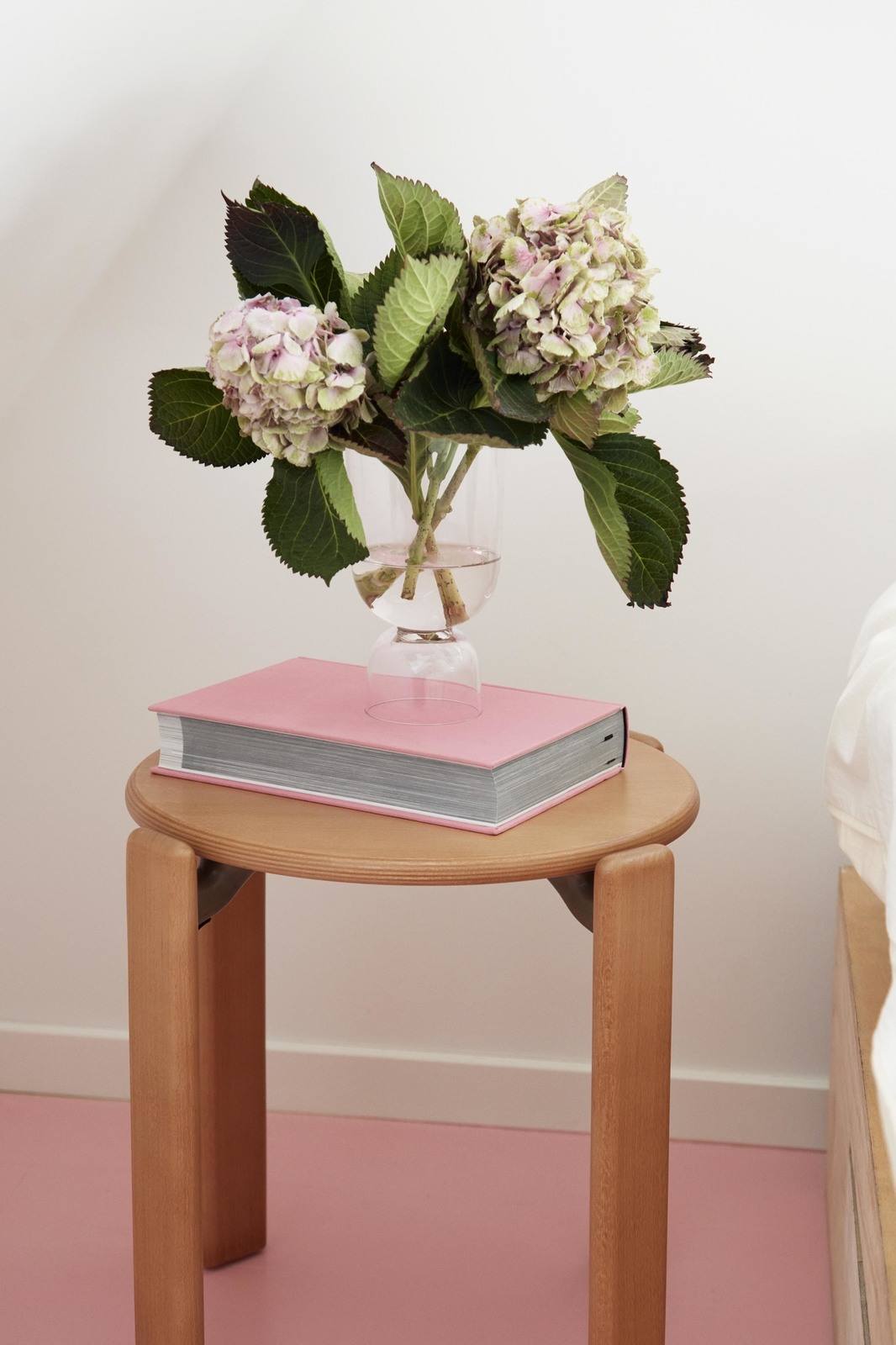 Hocker mit einem dicken Buch in Rosafarbenem Einband, darauf eine Glasvase mit zwei Hortensienblüten. 