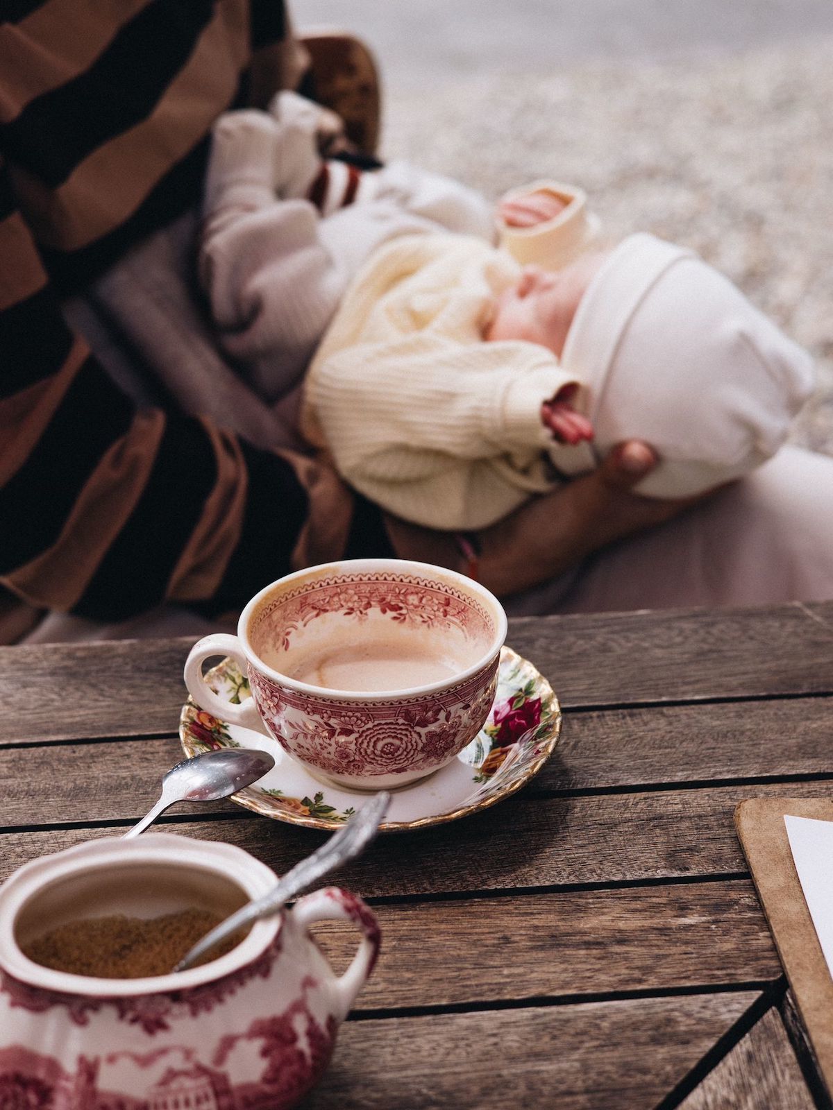 Im Vordergrund Kaffeetasse und Zuckerdose, im Hintergrund sitzt verschwommen eine Person, die ein Baby im Schoß hält.