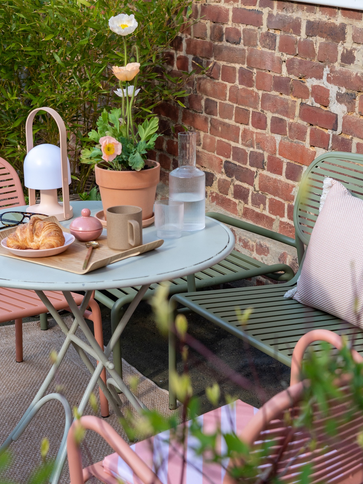 Sitzgruppe im Garten, auf dem Tisch steht ein Blumentopf mit Islandmohn, eine tragbare Leuchte, eine Wasserkaraffe und ein kleines Tablett mit Croissant und Tasse
