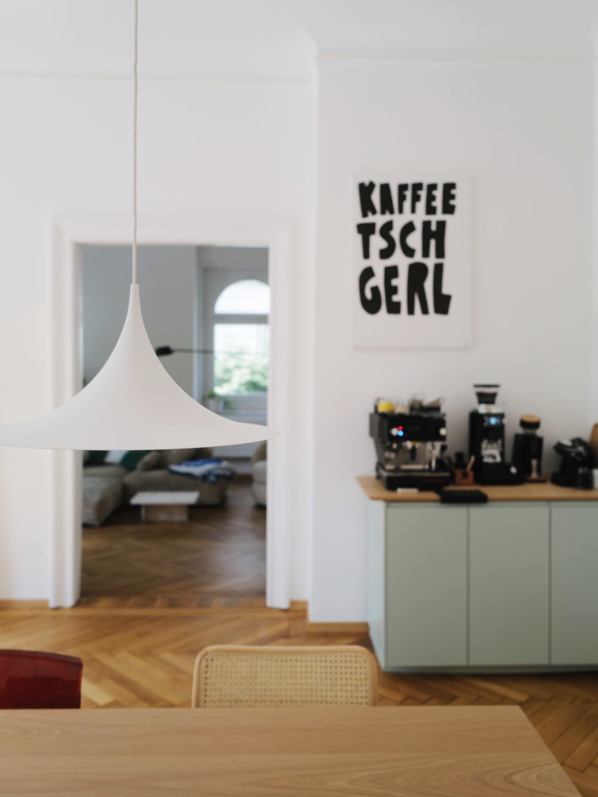 Eine weiße Pendelleuchte hängt über einem Esstisch, im Hintergrund steht eine Kaffeetheke und ein Poster mit dem Text "Kaffee Tschgerl" hängt darüber. 