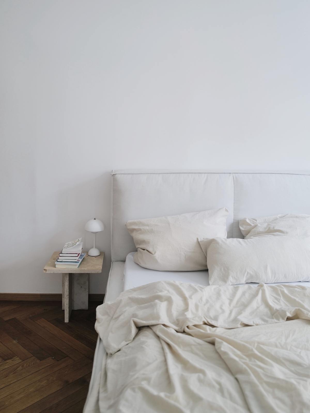 Bett mit gepolstertem Rahmen in Eierschalenfarben, dazu cremeweiße Bettwäsche und ein kleiner Beistelltisch aus hellem Naturstein. 