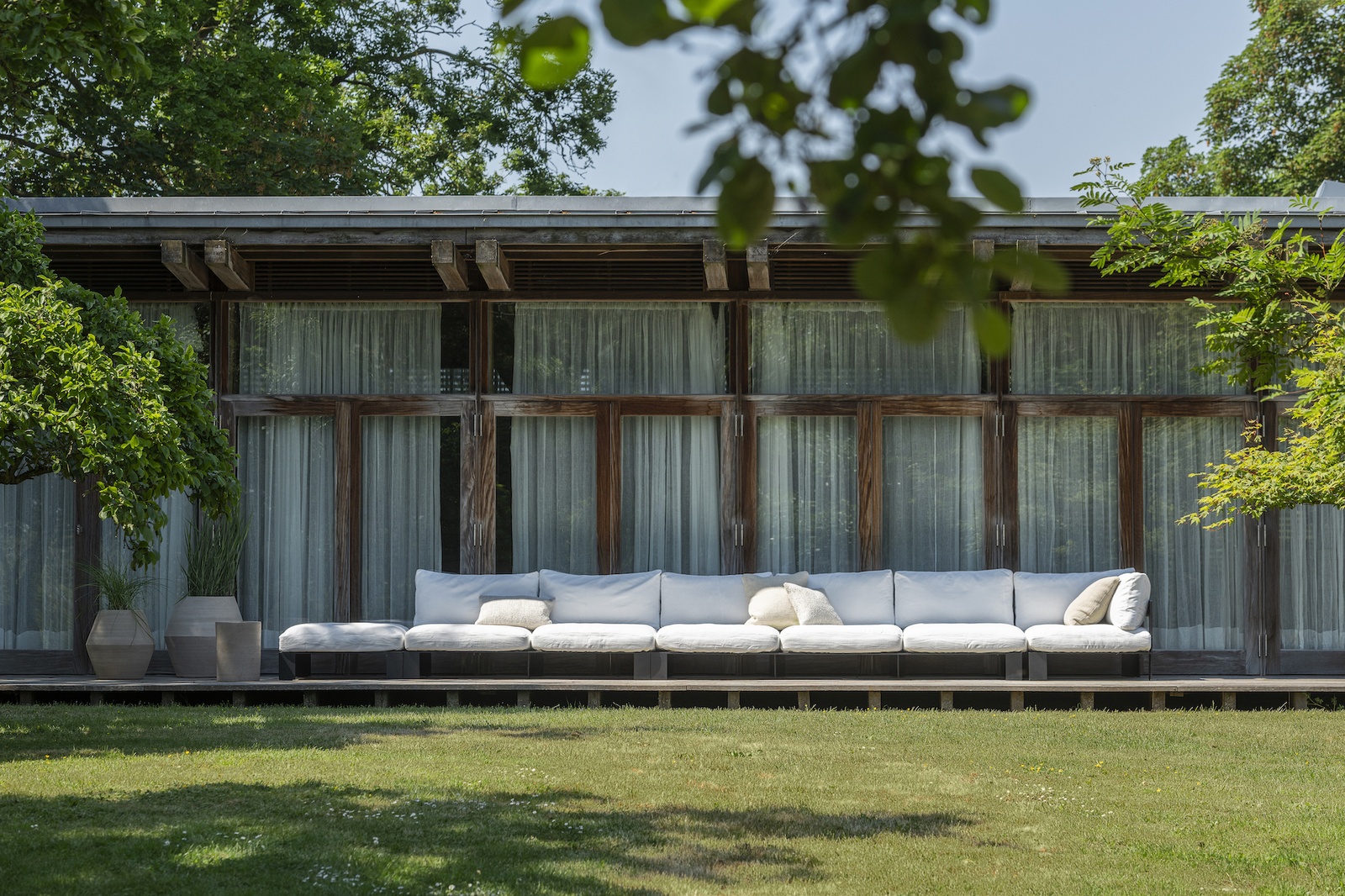 Glaspavillion mit Holzterrasse, auf der ein meterlanges Outdoor Loungesofa mit weißen bezügen steht.
